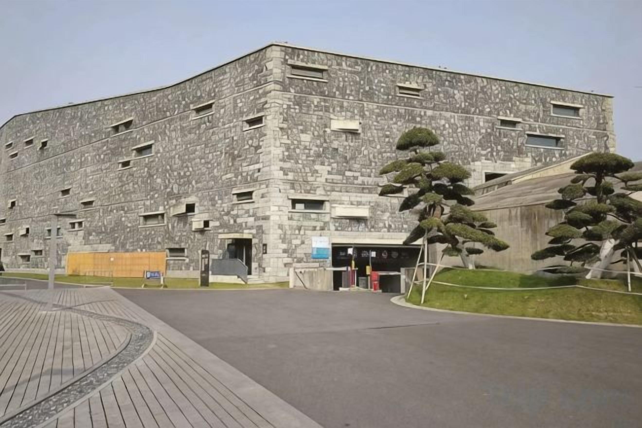A large, multi-story building with a stone facade, a paved entrance area, and a few trees.
