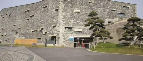 A large, multi-story building with a stone facade, a paved entrance area, and a few trees.