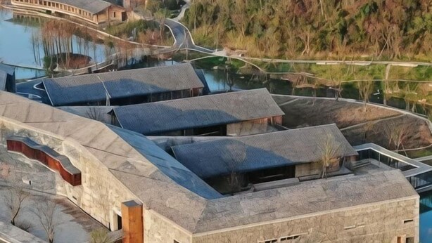 Un edificio moderno con techo curvo, piscina y un paisaje montañoso cubierto de bosque al fondo.