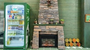Una máquina expendedora verde con diversas bebidas y aperitivos, una chimenea de piedra con una decoración de cabeza de ciervo, y una hilera de frutas coloridas a la derecha.