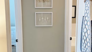 A hallway with beige walls, two framed artworks, a clock, and a decorative rope on a curtain. - Renovated 1st Floor 2 Bedroom Condo close to pool and beach with outdoor space. (Ocean City)