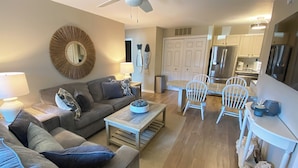 A modern living room with a grey sofa, a coffee table, and a dining area with white chairs. - Renovated 1st Floor 2 Bedroom Condo close to pool and beach with outdoor space. (Ocean City)