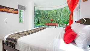 A bedroom with a large bed, a red canopy, a view of greenery outside, and a traditional artwork on the wall.