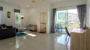 A spacious living room with a sofa, a glass table, and a wooden desk. There is a balcony with a view of greenery and a mountain.