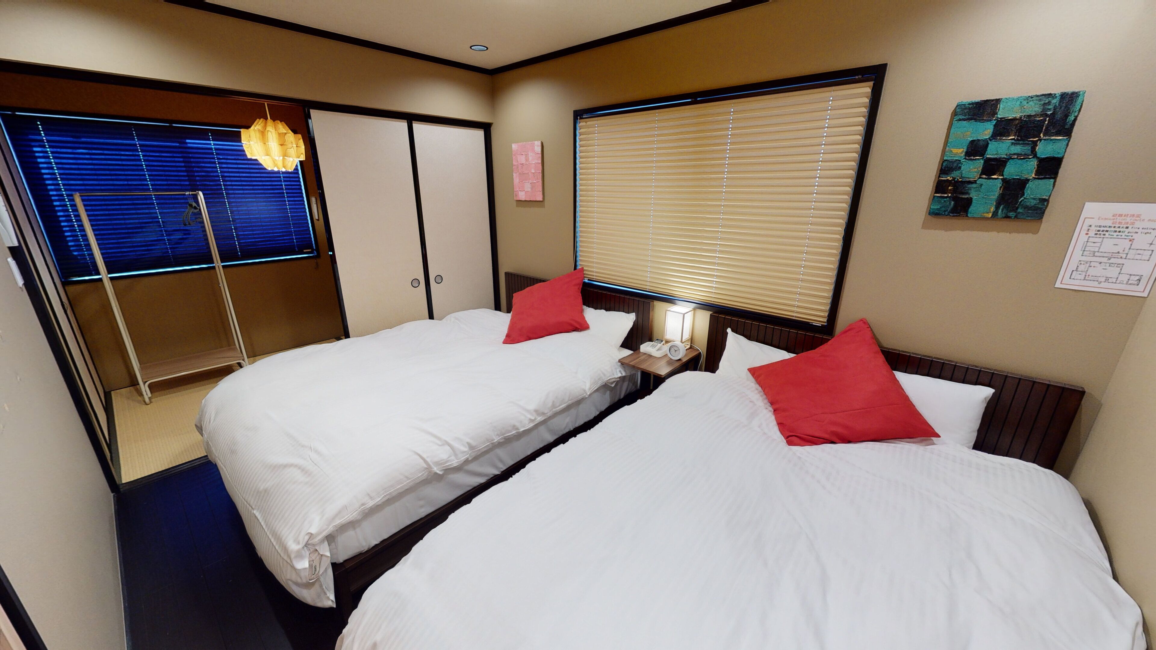 A hotel room with two beds, a wooden headboard, a bedside table with a lamp, a clock, and a window with blinds.