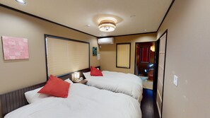 A hotel room with two beds, a wall-mounted air conditioner, a bedside table with a lamp, and a window with blinds.