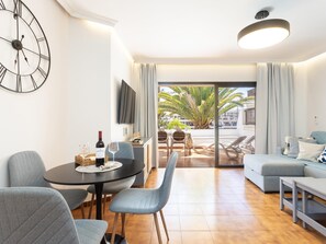 A modern living room with a dining area, a sofa set, a TV mounted on the wall, and a large window with a view of palm trees.