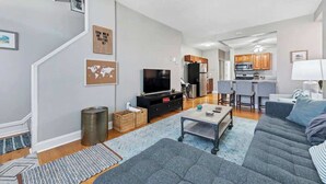 A modern living room with a grey sofa, a coffee table, and a TV. There is a kitchen in the background with wooden cabinets and a microwave. - 3 King Beds/3Baths Spacious by StayStLouis (St. Louis)