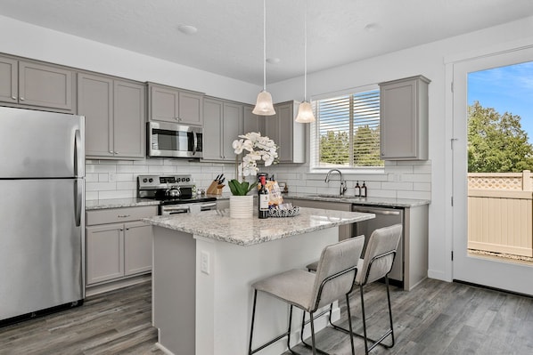 A modern kitchen with grey cabinets, stainless steel appliances, a granite countertop, and a large island with bar stools.