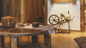A rustic wooden table with a woven basket, a small bowl, and a decorative cloth. In the background, there's a spinning wheel and a white wall with a wooden door.