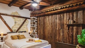 A rustic bedroom with a stone wall, wooden beams, a bed with a map, and a ceiling fan.