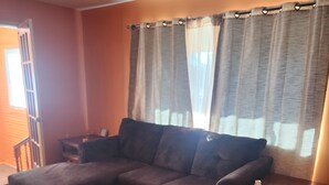 A living room with a sectional sofa, wooden flooring, a small table, and a window with curtains.