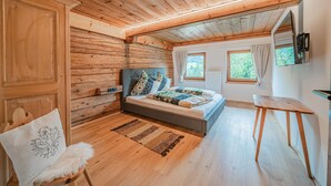 Dormitorio de una cabaña de madera con una cama, una silla, una mesita y una ventana con vistas a la vegetación.