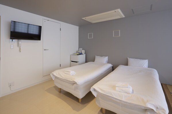 Habitación de hotel con dos camas individuales, un televisor montado en la pared, una mesita de noche pequeña con lámpara y una puerta.