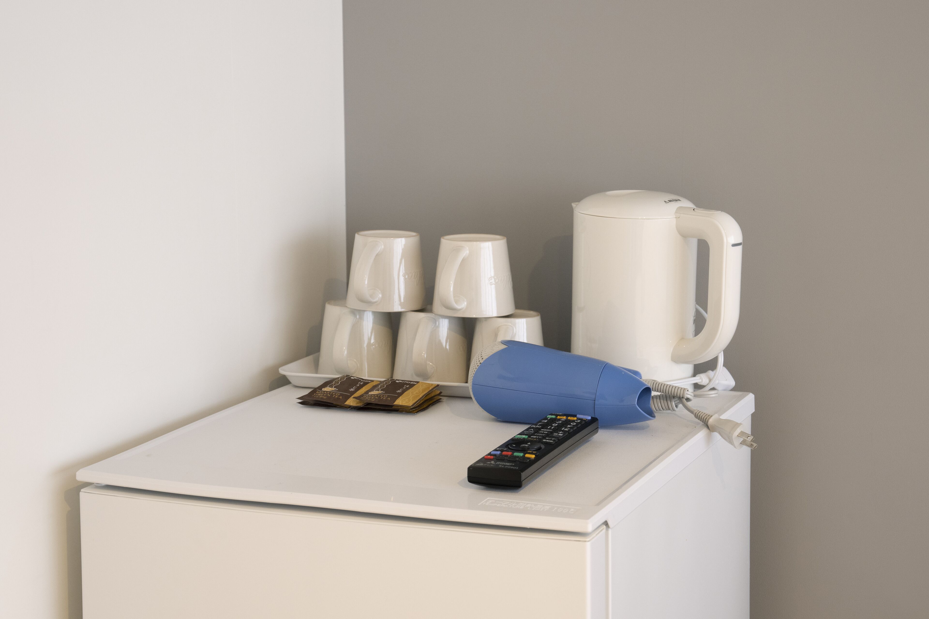 A white bedside table with a remote control, a blue pillow, a coffee maker, and a stack of white cups.