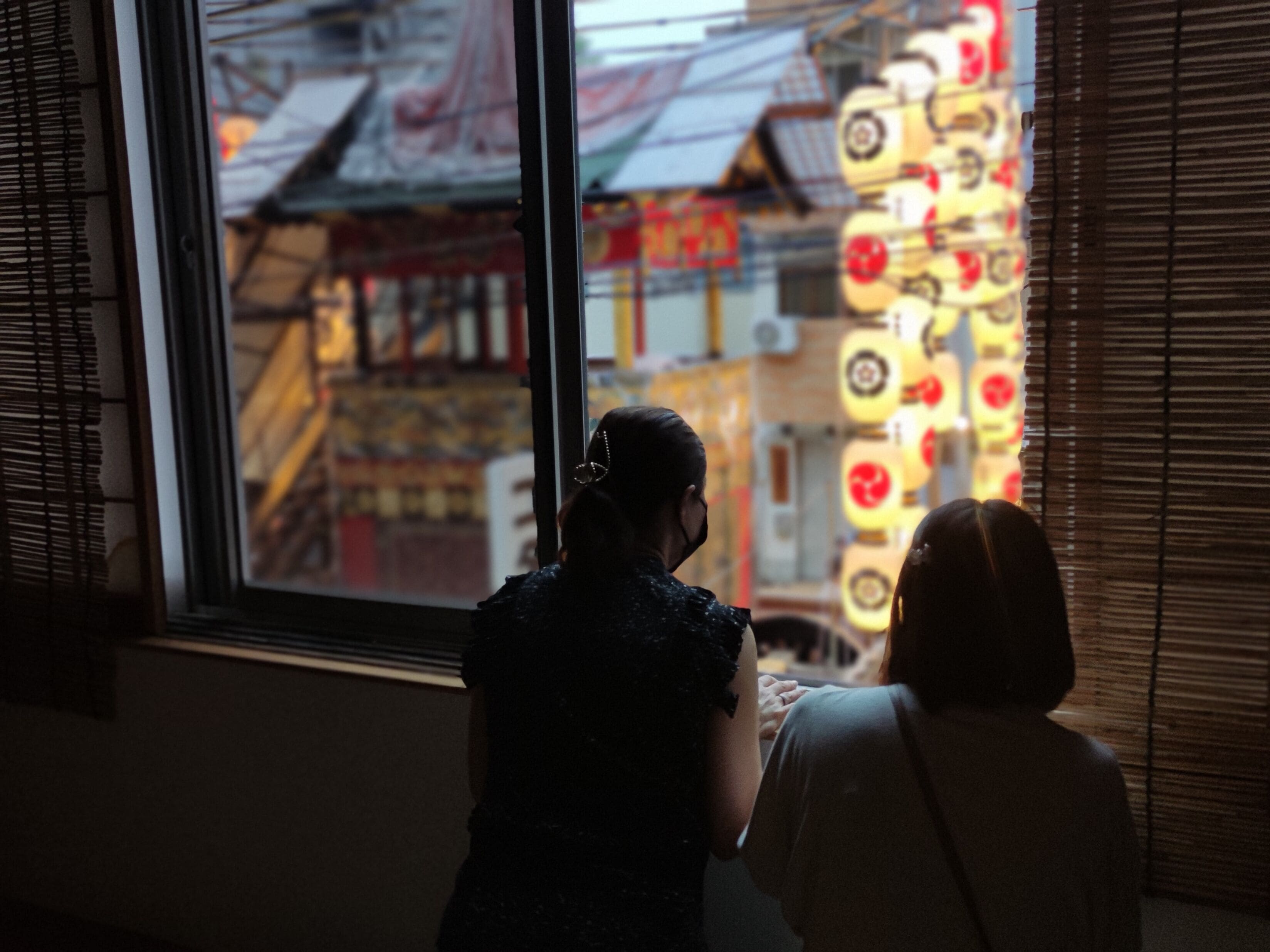 A view through a window with blinds, featuring a building with a traditional architectural style and a brightly lit sign with red and white colors.