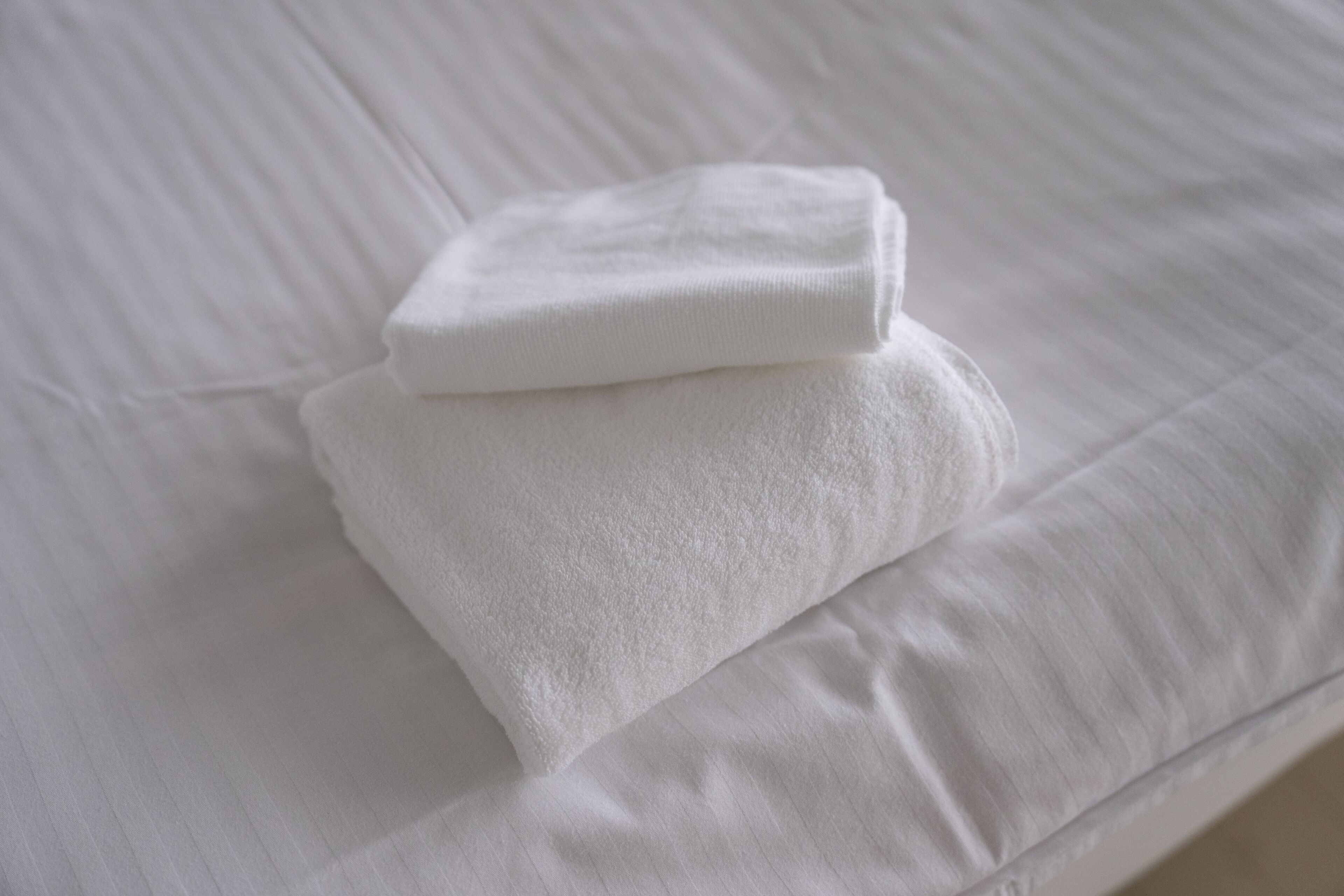 Une serviette blanche, soigneusement pliée, est posée sur un lit recouvert d’un drap blanc et lisse.