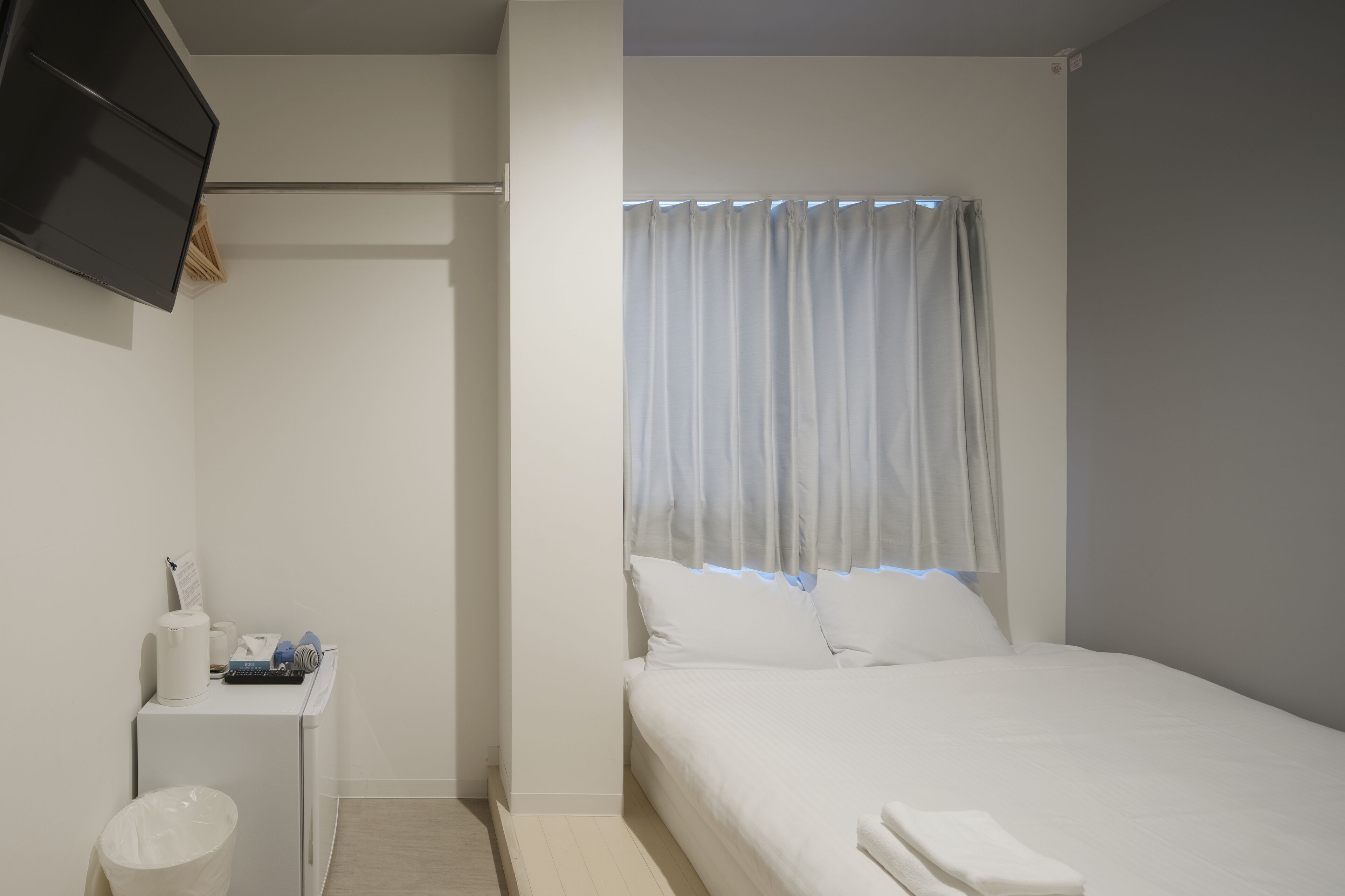 Et minimalistisk hotelværelse med en seng, et lille skrivebord med skraldespand, et fladskærms-TV monteret på væggen og et vindue med lette gardiner.
