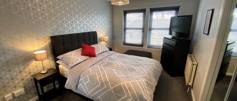A bedroom with a bed, bedside lamps, a dresser, a TV, and a window with blinds.