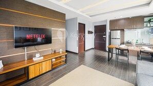 A modern living room with a flat-screen TV, wooden entertainment unit, and a dining area with a kitchen in the background. - PECAN by Kozystay (Jakarta)