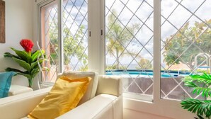 Ein Zimmer mit einer weißen Couch, einem gelben Kissen und Blick auf ein Schwimmbecken durch rautenförmige Fenster.