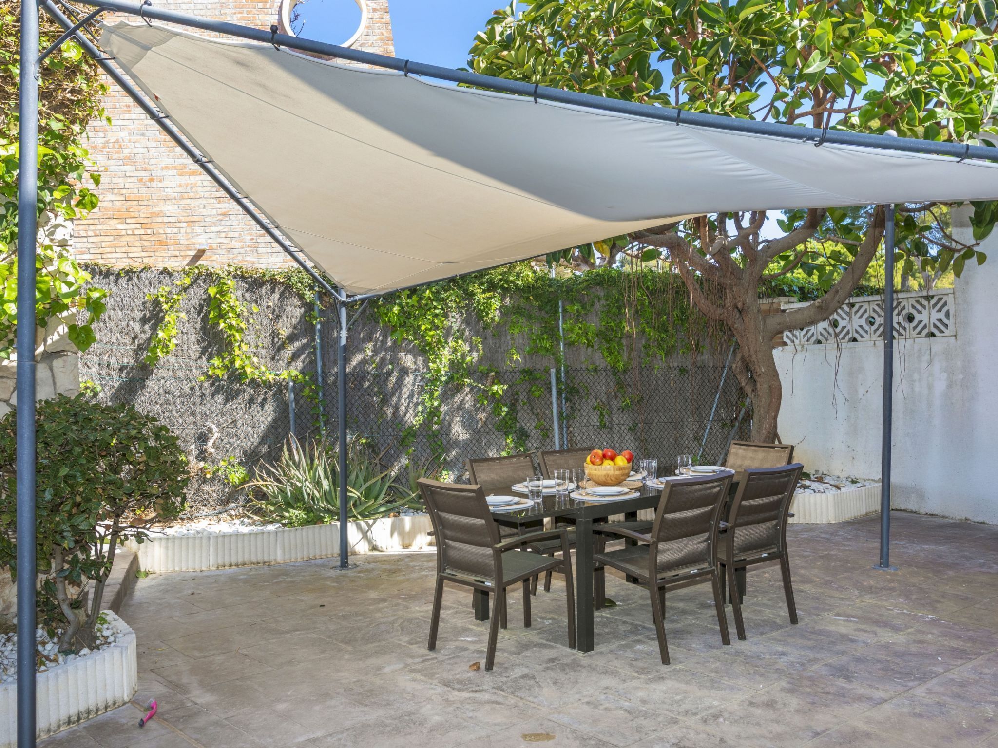 Une terrasse couverte avec une table dressée pour les repas, entourée de chaises, et un arbre qui offre de l’ombre.