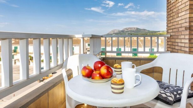 Un balcón con mesa dispuesta para el desayuno, que incluye manzanas y muffins, con vistas a un estacionamiento y montañas.