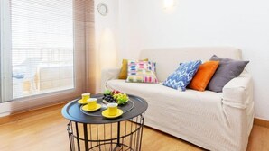 Una sala de estar acogedora con un sofá, cojines de colores, una pequeña mesa redonda con tazas y un frutero, y una ventana con persianas.