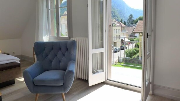 A room with a large window, a blue armchair, and a view of a town and mountains. - Henry's 300 by Interhome (Interlaken)