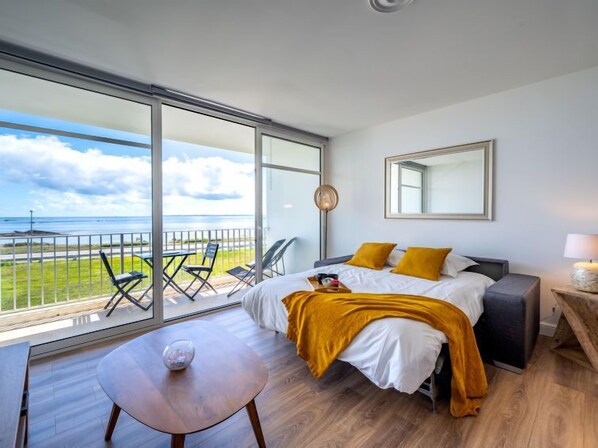 A modern hotel room with a large balcony, a bed with white bedding and mustard pillows, a round table with a decorative item, and a large mirror on the wall. - Résidence Les Trois Iles by Interhome (Quiberon)
