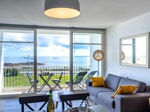 En moderne stue med en grå sofa, gule puder og et spisebord med stole. Et stort vindue giver udsigt over havet og en græsplæne.