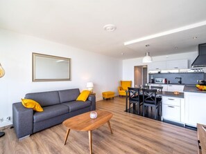 A modern living room with a grey sofa, wooden coffee table, and yellow cushions. A dining area with a black table and chairs is visible. The kitchen has white cabinets and a black range hood. - Résidence Les Trois Iles by Interhome (Quiberon)