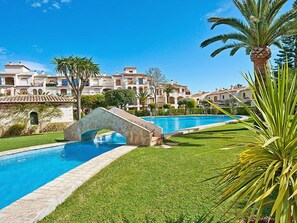A swimming pool with a stone bridge, surrounded by well-maintained grass and palm trees, with buildings in the background. - La Isla C12 by Interhome (Jávea)