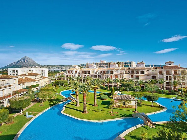 A resort with a large swimming pool, palm trees, and a mountain in the background. - La Isla C12 by Interhome (Jávea)
