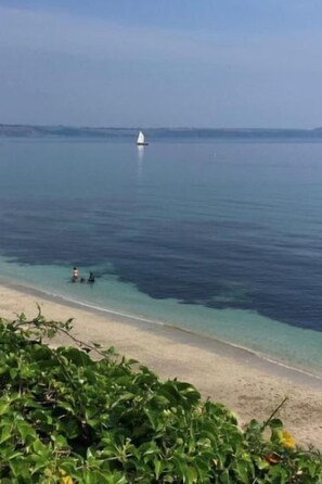 Une plage à l’eau turquoise et limpide, un voilier à l’horizon et quelques personnes près du rivage.