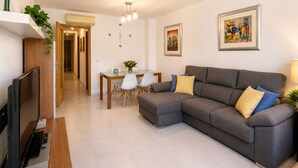 Un salon avec un canapé gris, un téléviseur à écran plat, une table à manger avec des chaises, et un couloir menant à une autre pièce.