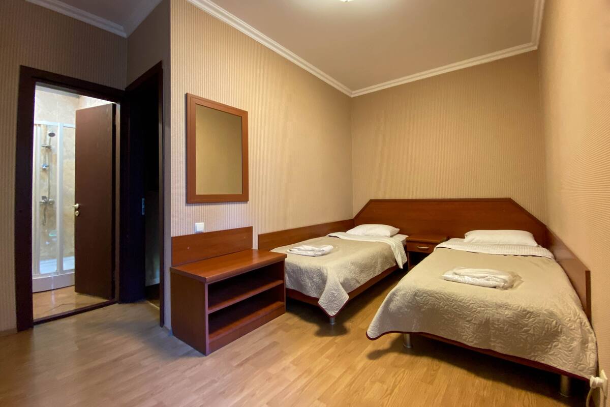 A hotel room with two single beds, a wooden bench, a large mirror, and a shower area.