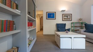 Un salón moderno con un sofá blanco, una mesa de centro y estanterías llenas de libros. Hay dos cuadros en la pared y una estantería de metal verde.