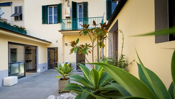Un patio con plantas en macetas, una mesa con mantel estampado y un edificio con contraventanas verdes y un balcón.