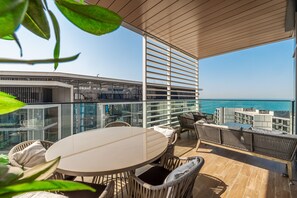 A modern outdoor balcony with a round table and wicker furniture, overlooking a cityscape and the sea.