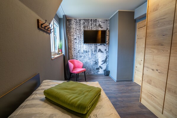 Une chambre d’hôtel avec un lit, une couverture verte, une chaise rose, un téléviseur fixé au mur et une armoire en bois.