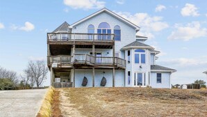 A large, two-story house with a balcony on the second floor, a small tower-like structure on the right, and a covered entryway. - 1525 - Pampa Hill - Poolside Corolla Getaway w/ Theater Room (Corolla)