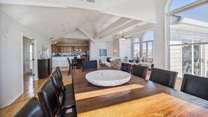A spacious dining area with a large wooden table and modern chairs, a kitchen in the background, and large windows offering a view of the city. - 1525 - Pampa Hill - Poolside Corolla Getaway w/ Theater Room (Corolla)