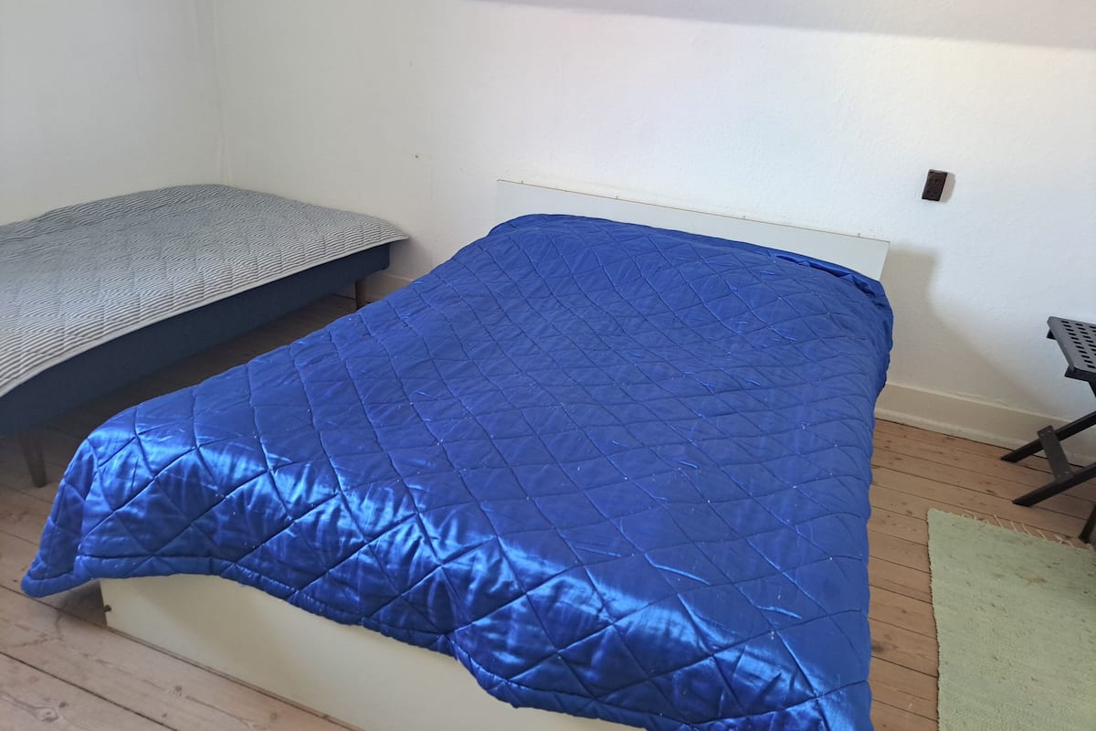A bed with a blue quilt, a grey patterned bedspread, and a green rug on a wooden floor.