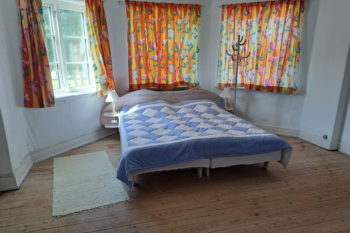 A bedroom with a wooden floor, a bed with a blue quilt, floral curtains, and a bedside lamp.
