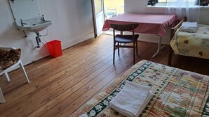 Une pièce avec un plancher en bois, un lit recouvert d’une courtepointe à motifs floraux, une table avec une nappe rouge, une chaise, un petit lavabo et une fenêtre avec des rideaux.