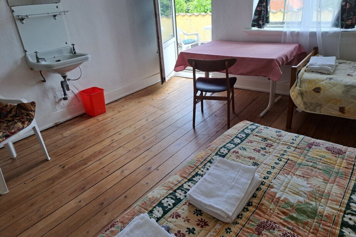 A room with a wooden floor, a bed with a floral quilt, a table with a red tablecloth, a chair, a small sink, and a window with curtains.