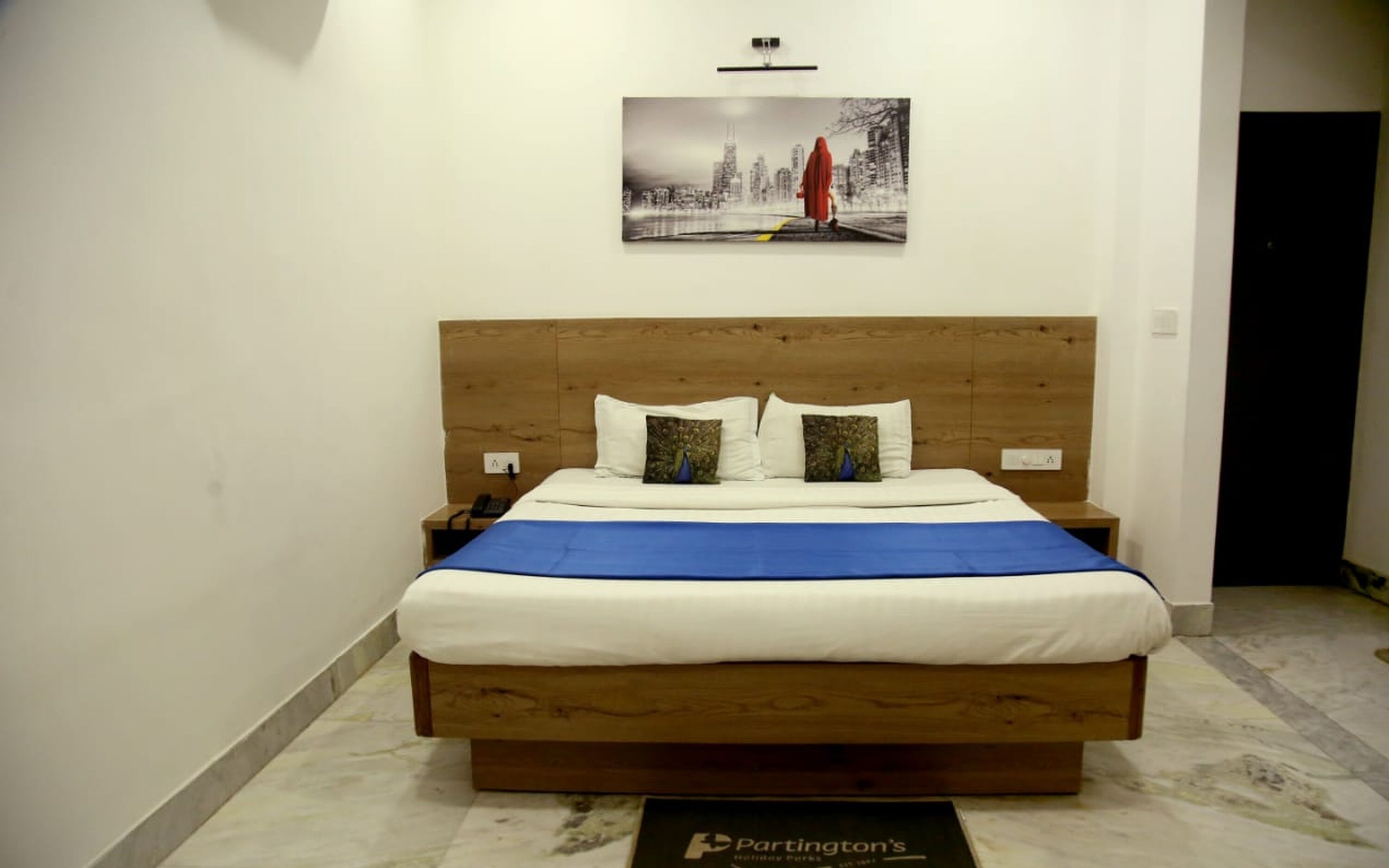 Une chambre d’hôtel avec un lit en bois, du linge de lit blanc et un tableau représentant un paysage urbain accroché au mur.