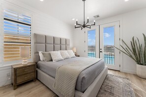 A bedroom with a large bed, a nightstand, a window with blinds, a chandelier, and a view of a pool through a glass door. - Sandy Beach Estate by RedAwning (St. George)