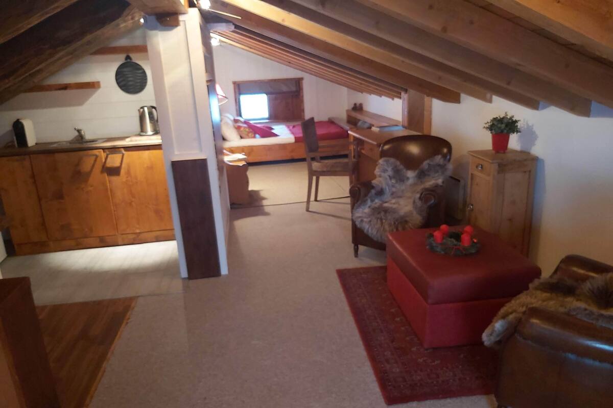 A cozy attic room with a wooden ceiling, a red ottoman, a chair with a fur throw, and a wooden cabinet.